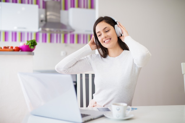 Jeune fille heureuse détendue avec un casque sur regarder un ordinateur portable boire du café et profiter de la musique tout en étant assis à la table de la cuisine.