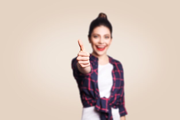 Jeune fille heureuse avec un style décontracté et des cheveux en chignon, le pouce en l'air, sur un mur blanc beige avec un espace de copie en regardant la caméra avec un sourire à pleines dents. se concentrer sur le doigt.