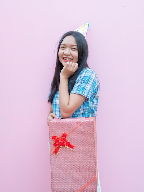 Photo une jeune fille heureuse tient une boîte à cadeaux et porte un chapeau de fête sur un fond rose.