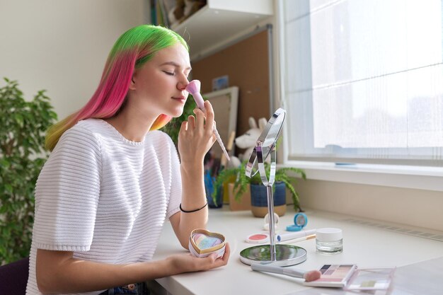 Jeune fille hipster aux cheveux colorés teints à la mode fait du maquillage, une adolescente assise à la maison à table près de la fenêtre avec une brosse et des cosmétiques décoratifs