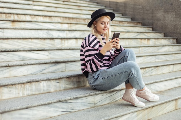 Une jeune fille hipster charmante avec un chapeau qui utilise son téléphone alors qu'elle est assise sur les escaliers à l'extérieur