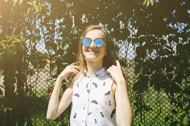 Jeune fille hipster en forme se tient près de la clôture des feuilles de lierre Femme en jupe bleue et chemisier sourit et profite du soleil Émotions drôles et heureuses en séance photo d'été