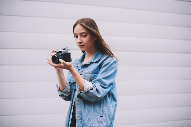 Jeune fille hipster vérifiant des photos à la caméra rétro tout en se tenant près du mur blanc à la mode