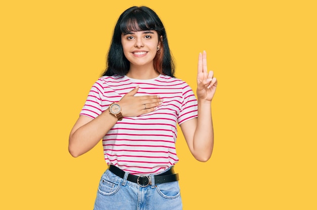 Jeune fille hispanique portant des vêtements décontractés souriant jurant avec la main sur la poitrine et les doigts vers le haut, faisant un serment de fidélité