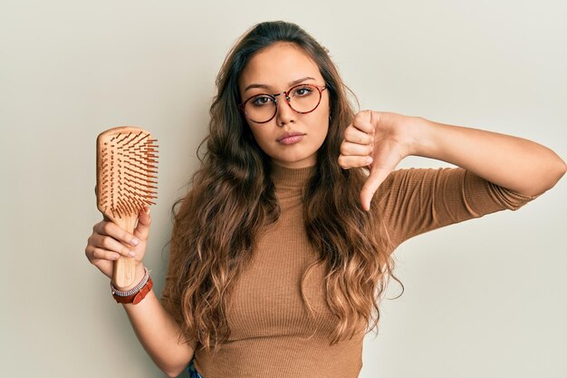Photo jeune fille hispanique tenant un peigne à cheveux avec un signe négatif de visage en colère montrant l'aversion avec le concept de rejet des pouces vers le bas