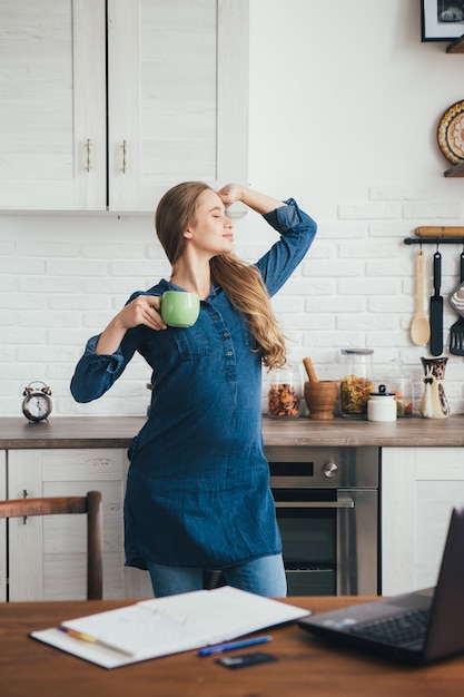 Jeune fille indépendante assez enceinte travaille à la maison dans la cuisine en mode auto-isolement en quarantaine et s'échauffe contre la fatigue