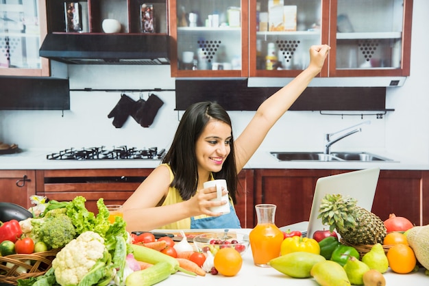 Jeune fille indienne attirante avec le tablier faisant cuire dans la cuisine avec la table pleine de fruits et légumes