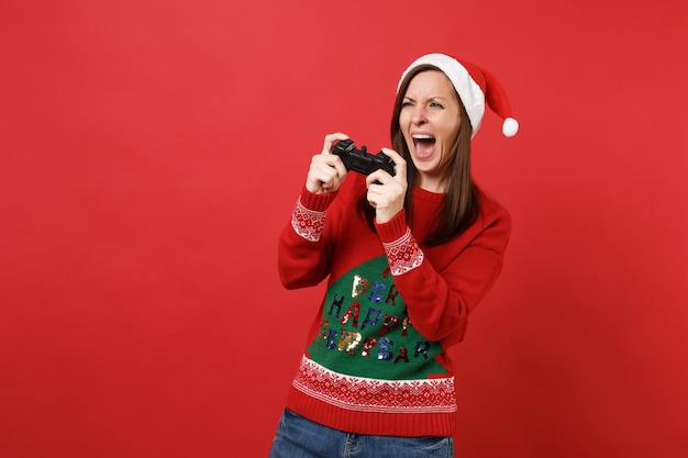 Jeune fille insatisfaite du Père Noël au chapeau de Noël criant, jouant à un jeu vidéo avec une manette isolée sur fond rouge vif. Bonne année 2019 concept de fête de vacances célébration. Maquette de l'espace de copie.