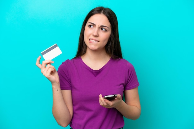 Jeune fille isolée sur fond bleu achetant avec le mobile avec une carte de crédit en pensant