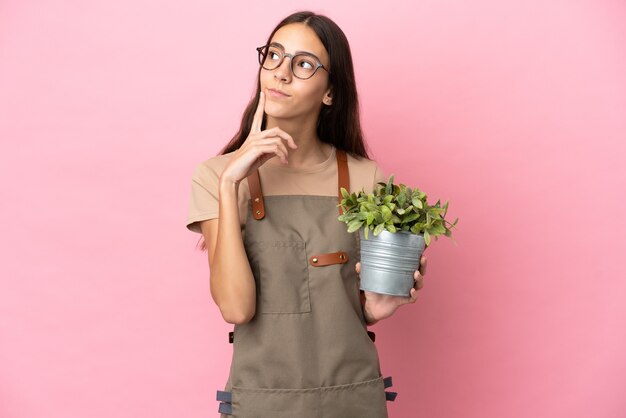 Jeune fille de jardinier tenant une plante isolée sur fond rose ayant des doutes tout en levant les yeux