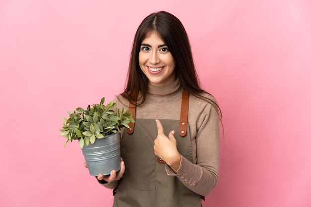 Jeune fille de jardinier tenant une plante isolée sur fond rose avec une expression faciale surprise