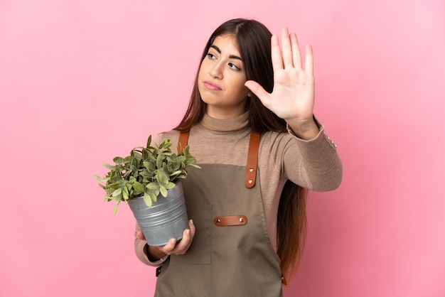 Jeune fille de jardinier tenant une plante isolée sur fond rose faisant le geste d'arrêt et déçu