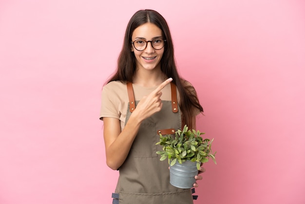Jeune fille de jardinier tenant une plante isolée sur fond rose pointant vers le côté pour présenter un produit