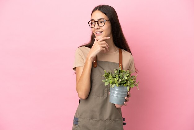 Jeune fille de jardinier tenant une plante isolée sur fond rose à la recherche sur le côté et souriant