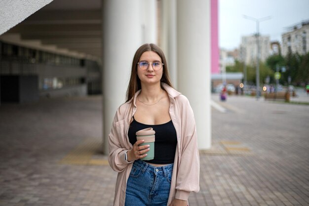 une jeune fille en jeans et une chemise avec une tasse de café dans les mains se tient dans la rue