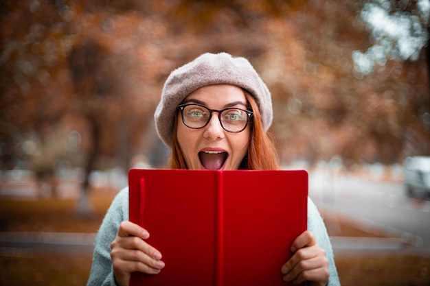 Jeune fille joyeuse émotionnelle dans un béret et un pull chaud tient un cahier rouge dans le parc en automne