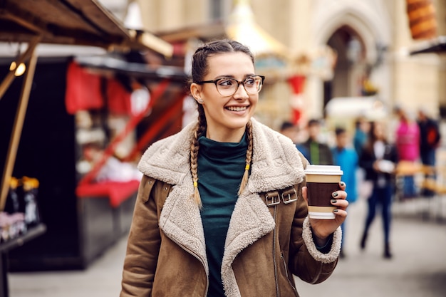 Jeune fille joyeuse marchant dans la rue du centre-ville, tenant une tasse jetable avec du café frais et profitant du temps libre le week-end.