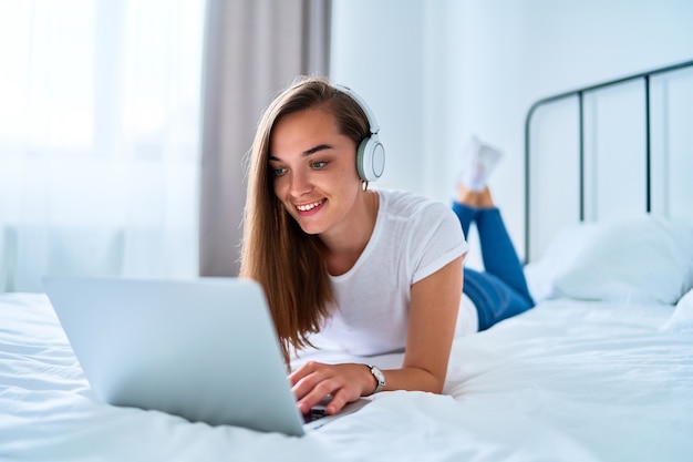 Jeune fille joyeuse souriante et joyeuse du millénaire allongée sur un lit et utilisant un ordinateur portable avec un casque sans fil pour regarder des vidéos en ligne, apprendre des cours de langue, des réseaux sociaux et des appels vidéo à la maison