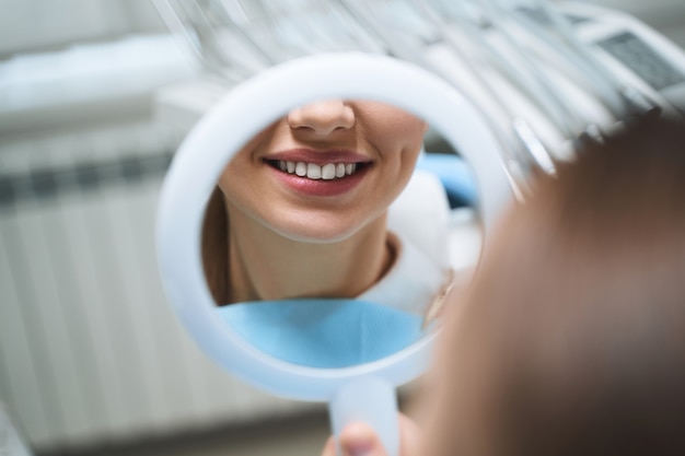 Une jeune fille joyeuse visite une clinique dentaire et regarde le reflet de son sourire après le traitement