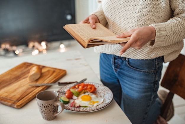 La jeune fille lit un vieux livre au petit déjeuner. Gros plan sur les mains et le réglage de la table. Style campagnard. Oeufs brouillés et légumes frais et lecture d'un livre intéressant le matin