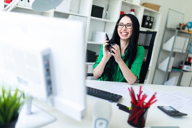 Une jeune fille avec des lunettes au bureau tient un téléphone dans ses mains et rit.