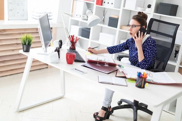Une jeune fille à lunettes est assise dans le bureau d'un bureau d'ordinateur et parle au téléphone.