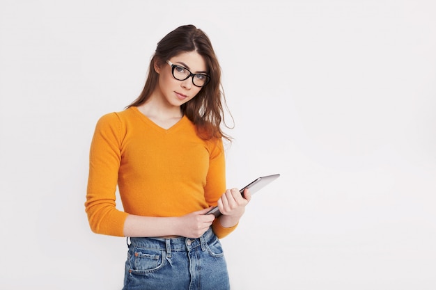 Une jeune fille à lunettes et un pull jaune tient une tablette et sourit. Portrait d'une jeune fille sur fond clair avec un espace pour le texte