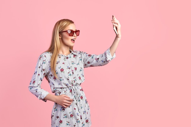 Jeune fille à lunettes de soleil portant une robe à fleurs, chapeau de paille, isolé sur fond rose.