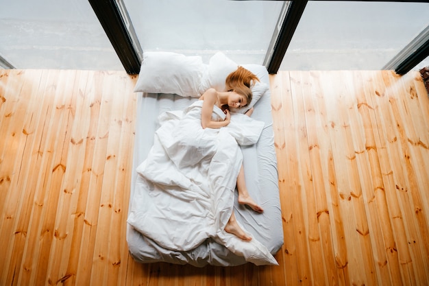 Jeune fille maigre pieds nus belle couchée dans un lit blanc avec couverture et oreillers et dormir. Le matin, détendez-vous au canapé. Gingembre, femme, reposer, dans, studio, apartmment, à, plancher bois, et, énorme, fenêtres