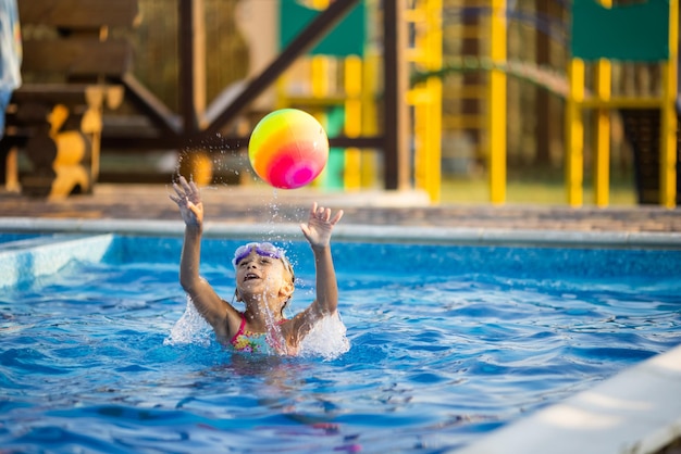 Une jeune fille en maillot de bain léopard brillant, nage avec un ballon gonflable de couleur vive dans une piscine d'un bleu profond avec de l'eau claire et transparente lors d'une chaude soirée d'été ensoleillée