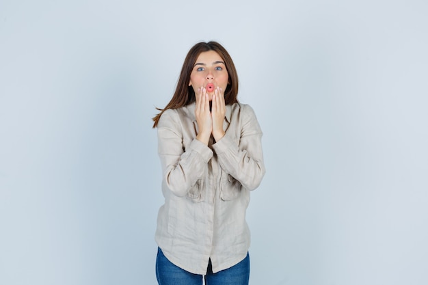 Jeune fille avec les mains près de la bouche en chemise beige, jeans et l'air surpris. vue de face.