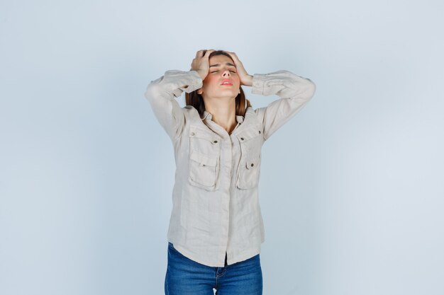 Jeune fille avec les mains sur la tête en chemise beige, jeans et semblant agitée, vue de face.