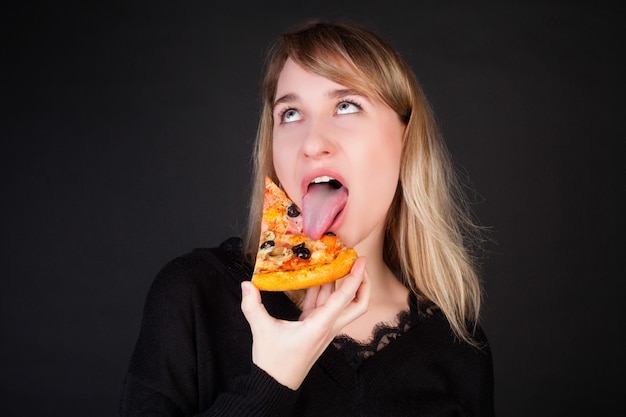 La jeune fille mange une tranche de pizza et fait une grimace, sur fond noir.