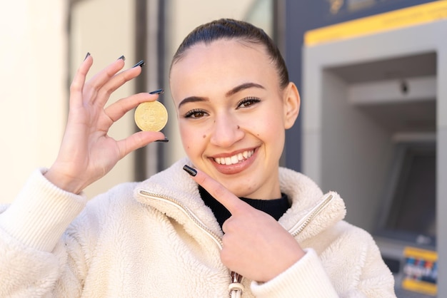 Jeune fille marocaine à l'extérieur tenant un Bitcoin et le pointant