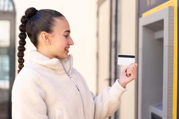 Photo jeune fille marocaine tenant une carte de crédit à l'extérieur avec une expression heureuse