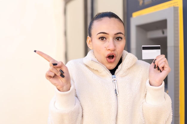 Jeune fille marocaine tenant une carte de crédit à l'extérieur surprise et pointant du côté