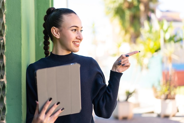 Jeune fille marocaine tenant une tablette à l'extérieur pointant vers le côté pour présenter un produit