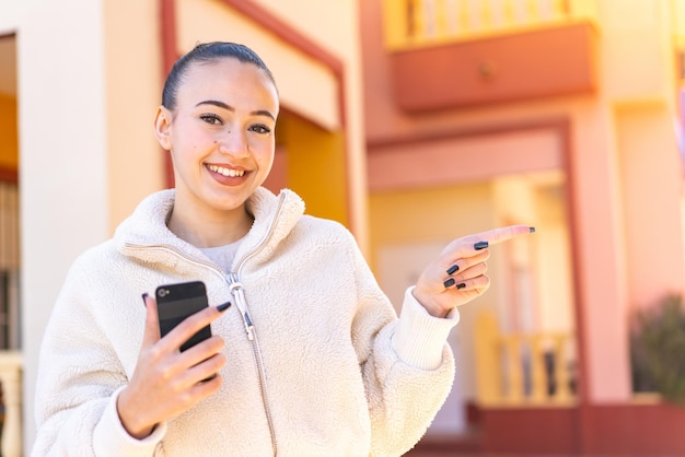 Jeune fille marocaine utilisant un téléphone portable à l'extérieur pointant vers le côté pour présenter un produit