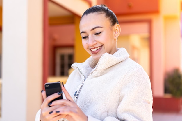 Jeune fille marocaine utilisant un téléphone portable à l'extérieur souriant beaucoup