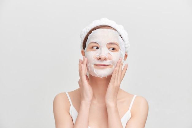 Jeune fille avec un masque facial regardant la caméra sur fond blanc. Procédure cosmétique. Spa de beauté et cosmétologie.