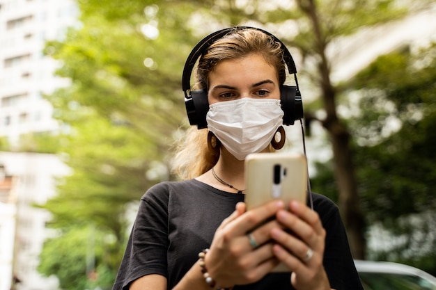 Jeune fille avec un masque de protection et des écouteurs pour écouter de la musique à l'aide de son téléphone portable dans la rue