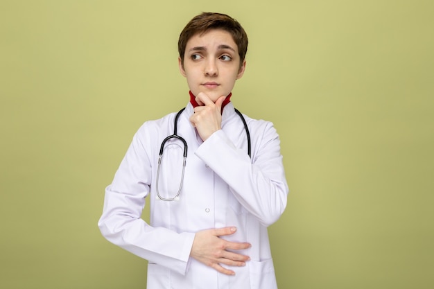 Jeune fille médecin en blouse blanche avec stéthoscope autour du cou à côté avec la main sur son menton pensant debout sur le vert