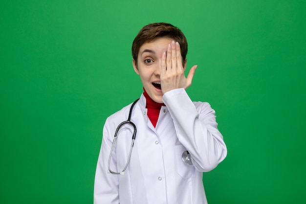 Jeune fille médecin en blouse blanche avec stéthoscope autour du cou heureux et positif couvrant un œil avec la main debout sur le mur vert