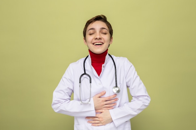 Jeune fille médecin en blouse blanche avec stéthoscope autour du cou souriant joyeusement tenant les mains sur sa poitrine ressentant des émotions positives debout sur un mur vert