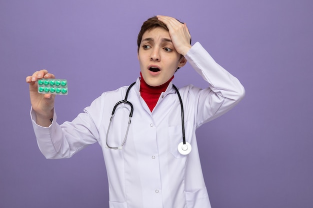 Jeune fille médecin en blouse blanche avec stéthoscope autour du cou tenant un blister avec des pilules le regardant confus et très anxieux debout sur un mur violet