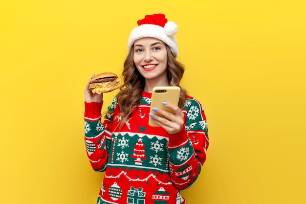 Une jeune fille mignonne en pull de Noël et un chapeau de Père Noël tient un grand cheeseburger et utilise un smartphone