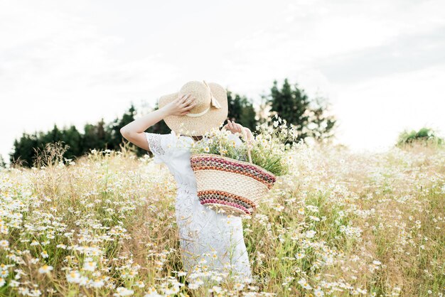 Jeune fille mignonne, tendre rêveuse amoureuse, dans un champ de marguerites. en robe et chapeau en osier. été chaud et ensoleillé, coucher de soleil dans le village. concept de liberté et style de vie