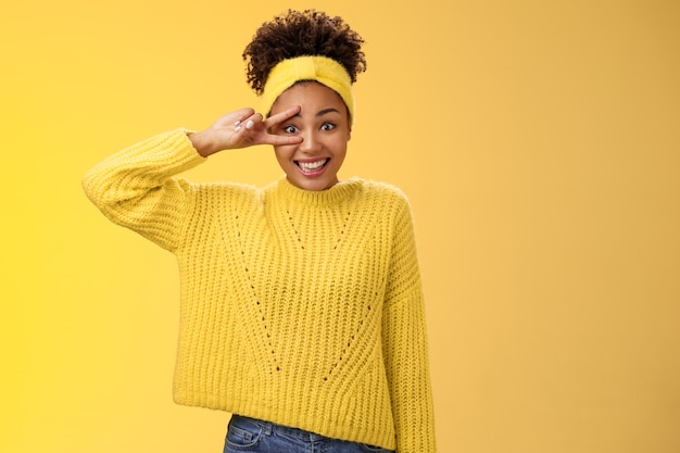 Une jeune fille millénaire maladroite et maladroite, idiote, rougissant, peu confiante, photographiant souriant, signe de victoire pour la paix près de l'œil mauvais en posant, debout sur fond jaune amical. Espace de copie