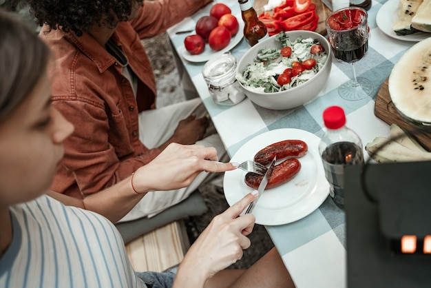 Jeune fille millénaire mangeant des saucisses grillées au glamping, assise à la table. Joyeux millénaires campant à un pique-nique en plein air sous des ampoules. Passer du temps avec des amis à l'extérieur, barbecue