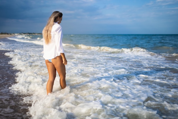 Une jeune fille mince dans un maillot de bain bleu doux et une chemise blanche, se promène le long d'une large plage de sable près de la mer bleue avec des vagues blanches